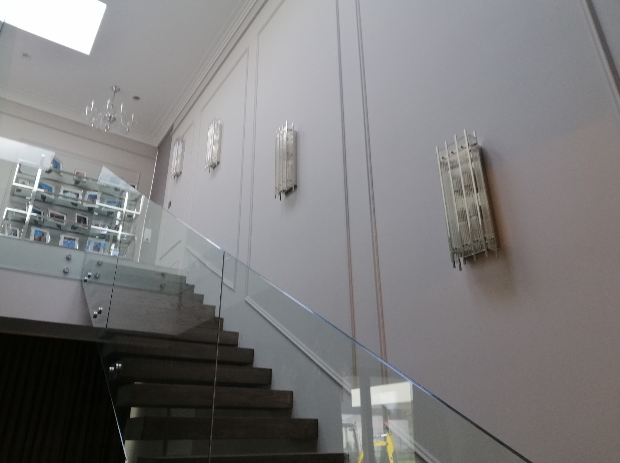 Eleganckie wykończenie ściany z panelami i oświetleniem schodów z balustradą ze szkła. Widoczny fragment schodów i żyrandol w tle. Stonowana kolorystyka.