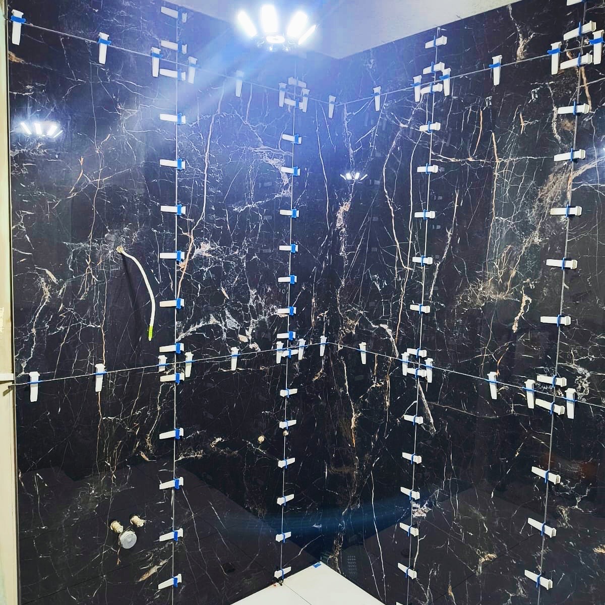 Montaż czarnych płytek imitujących marmur w łazience z użyciem systemu poziomowania. Widoczne białe klipsy i kliny. Oświetlenie sufitowe.