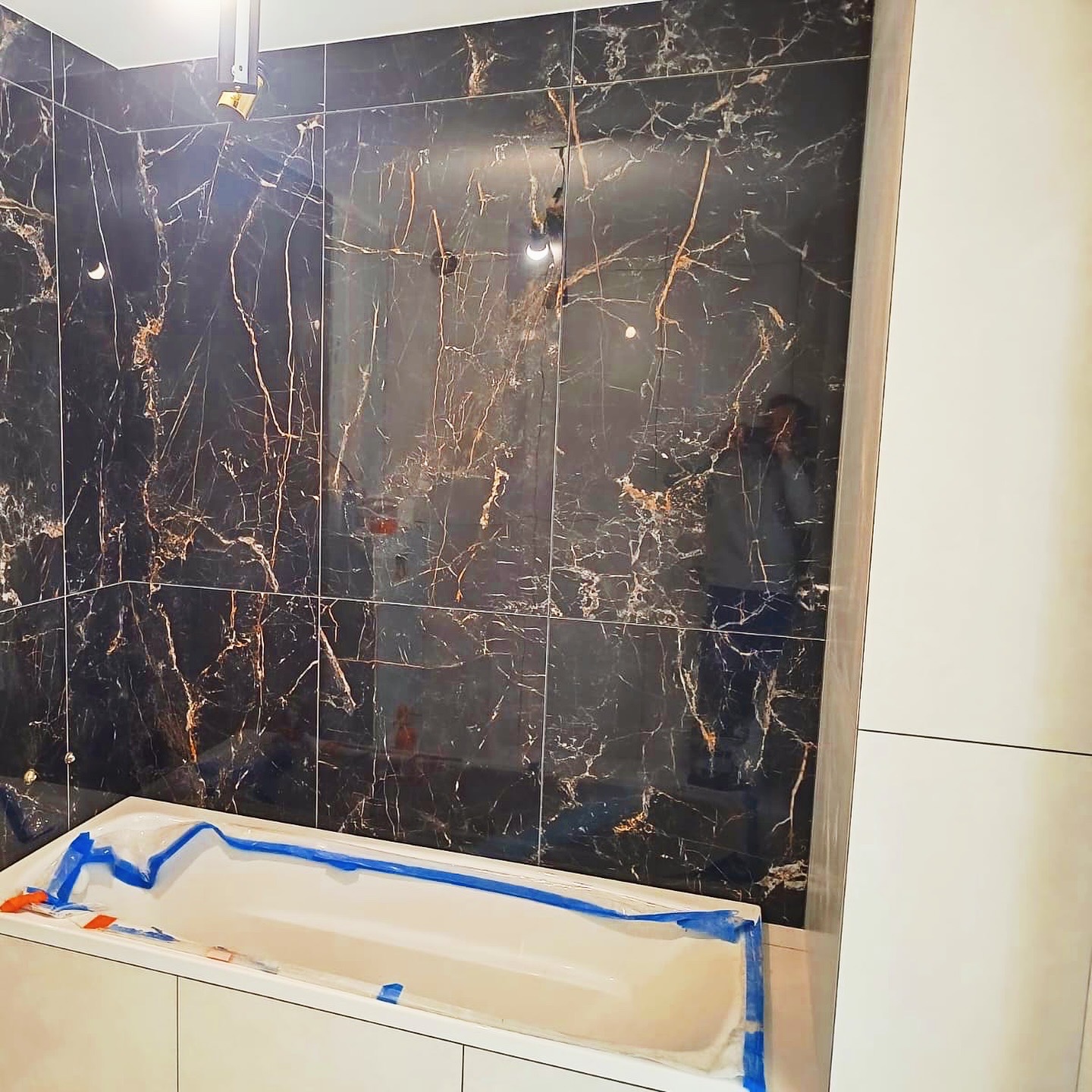 Czarne płytki imitujące marmur z złotymi żyłami na ścianie łazienki, zabezpieczona wanna w folii. Nowoczesne wykończenie w trakcie prac.