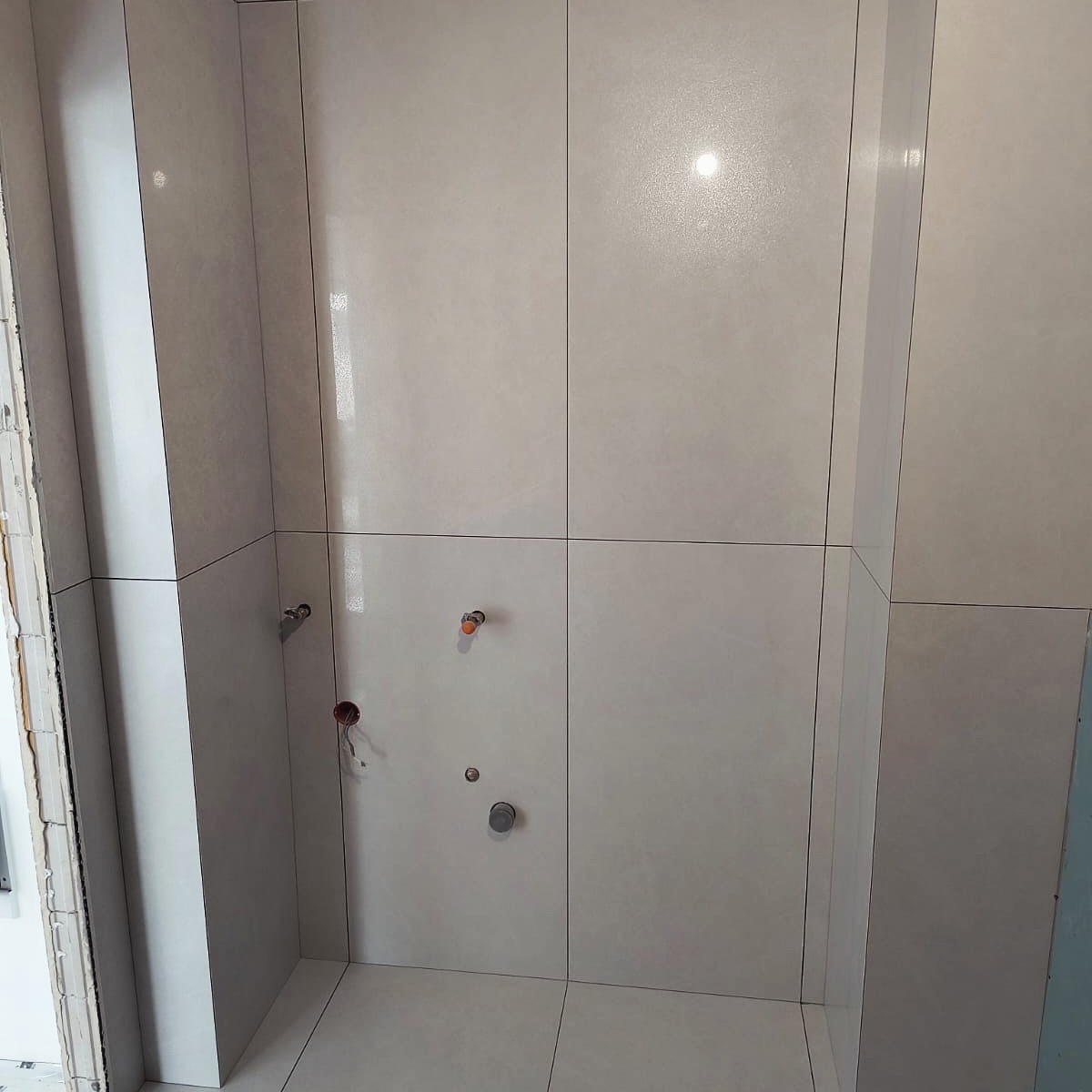 Wykończona łazienka z jasnymi, błyszczącymi płytkami ceramicznymi. Widoczne przyłącza wodne i kanalizacyjne przed montażem armatury. Minimalistyczny design.