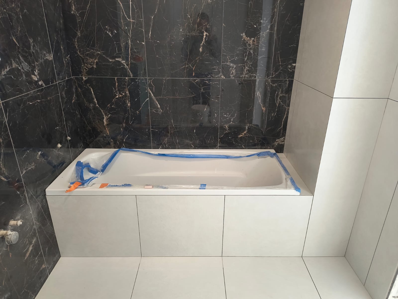 Biała wanna obudowana płytkami, zabezpieczona folią. Ściany wyłożone czarnymi płytkami imitującymi marmur i jasnymi, pionowymi płytkami. Wykończenie łazienki w trakcie.
