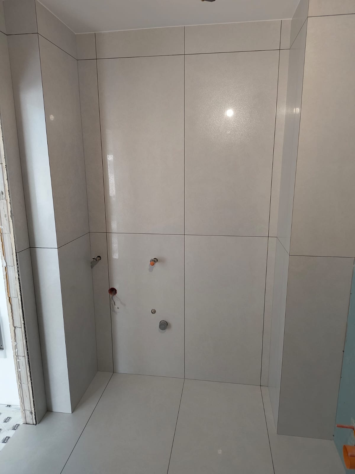 Wykończona łazienka z jasnymi, kwadratowymi płytkami na ścianach i podłodze. Widoczne przyłącza wodne i kanalizacyjne. Minimalistyczny design, czyste linie.