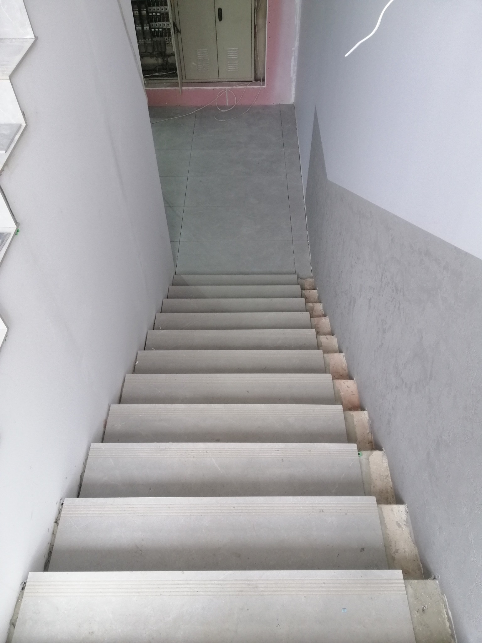 Wykończone schody w szarościach, widok z góry. Ściany pomalowane na szaro, płytki na schodach w odcieniach szarości. Widoczna szafka elektryczna w tle.