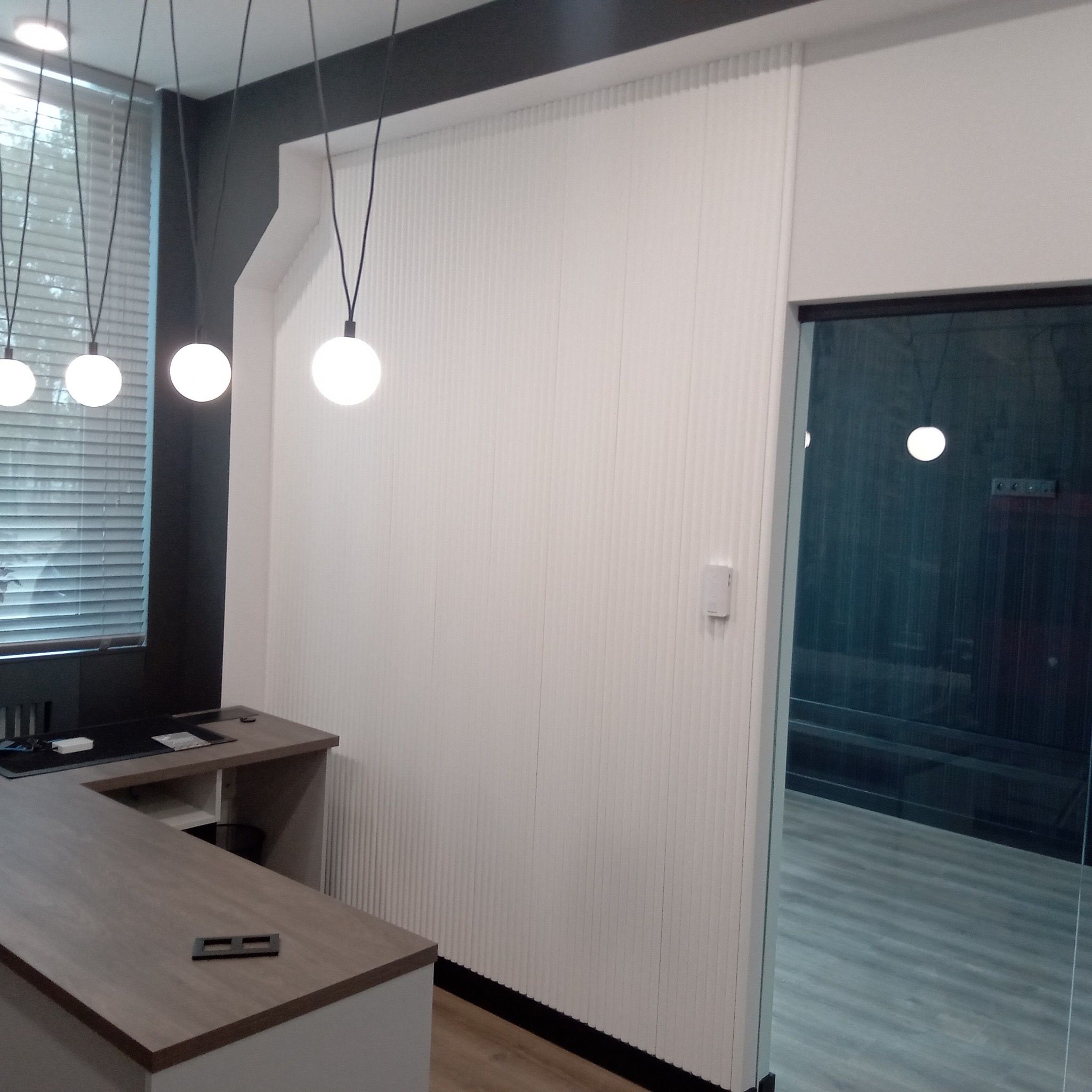 Biała ściana z pionowymi panelami dekoracyjnymi w nowoczesnym biurze, widoczne lampy wiszące i fragment biurka. Minimalistyczny design z elementami czerni.