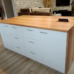 Meble pod każdy wymiar - Wyspa kuchenna z drewnianym blatem i białymi frontami szuflad, z wbudowanym gniazdkiem elektrycznym. Widoczne elementy wystroju wnętrza.
