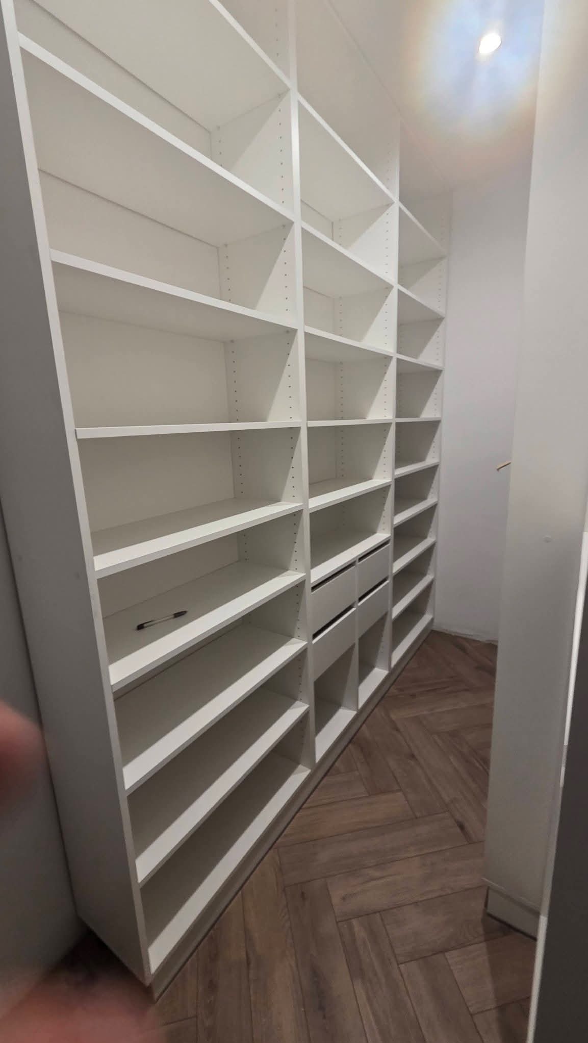Biała, zabudowana szafa wnękowa z półkami i szufladami, widok z dołu, na podłodze parkiet w jodełkę. Na jednej z półek leży długopis.