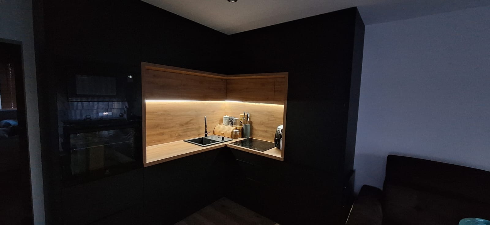 Nowoczesna, czarna kuchnia z drewnianym blatem i oświetleniem LED. Widoczny zlew, płyta indukcyjna i akcesoria kuchenne. Minimalistyczny design.