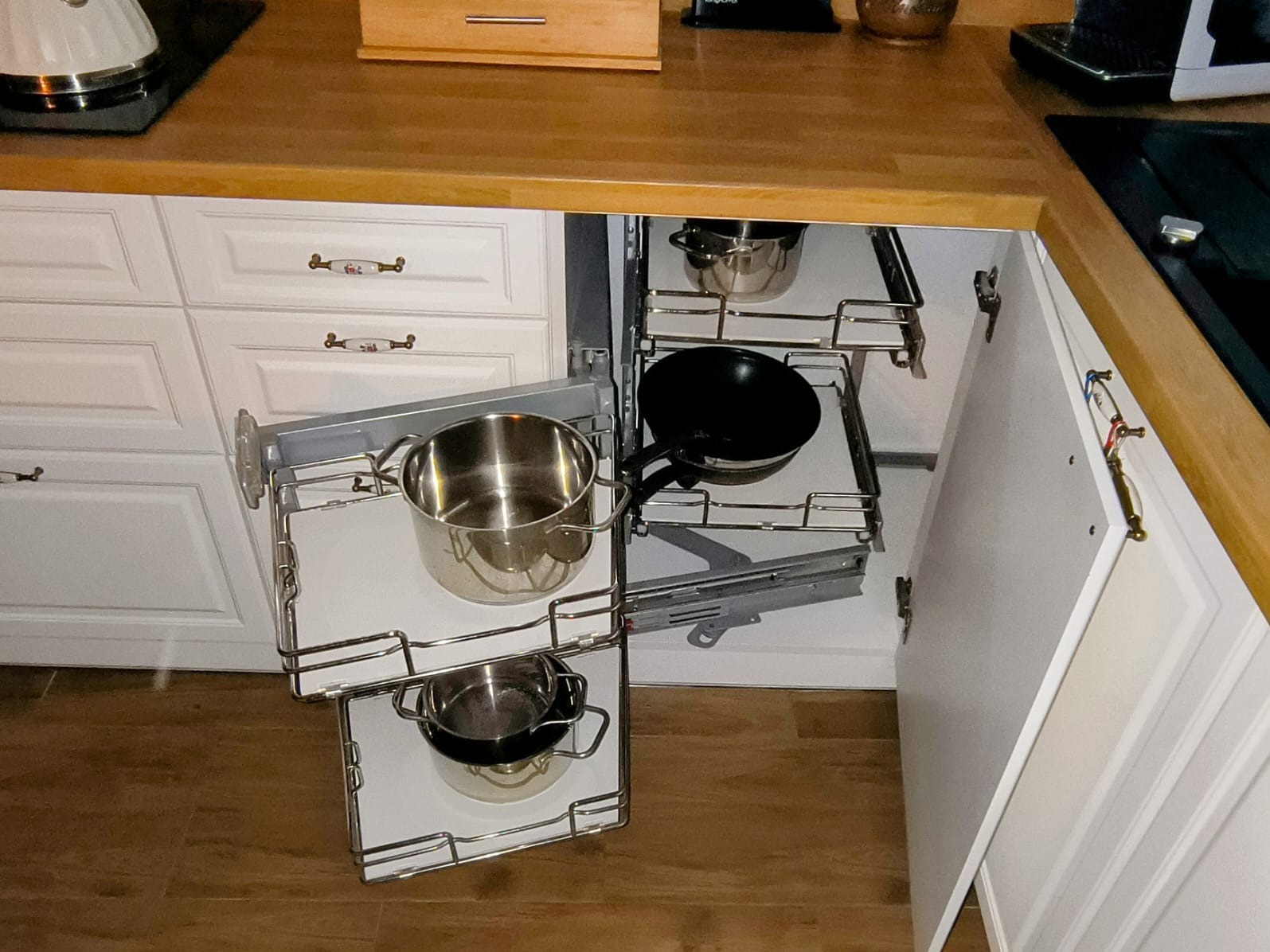 Wysuwana szafka narożna w kuchni z białymi frontami i drewnianym blatem, wyposażona w metalowe półki z garnkami i patelnią. Funkcjonalne rozwiązanie do przechowywania.