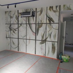 Usługi remontowo-budowlane - Ściana z dekoracyjną tapetą w motywy roślinne, podzielona ciemnymi listwami. Widoczne oświetlenie sufitowe i fragment klimatyzacji. Podłoga w trakcie prac wykończeniowych.