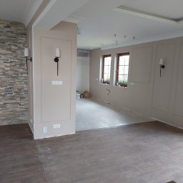 Usługi remontowo-budowlane - Jasne wnętrze po remoncie: brązowe panele podłogowe, beżowe ściany z dekoracyjnymi listwami, kamienna ściana, nowoczesne lampy ścienne. Widok na dalsze pomieszczenia.