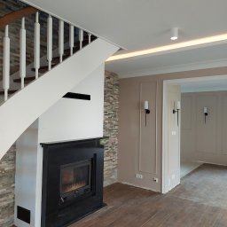 Usługi remontowo-budowlane - Salon po remoncie: drewnopodobna podłoga, kominek z czarną obudową i kamienną ścianą, białe schody, beżowe ściany z dekoracyjnymi listwami i czarnymi kinkietami.