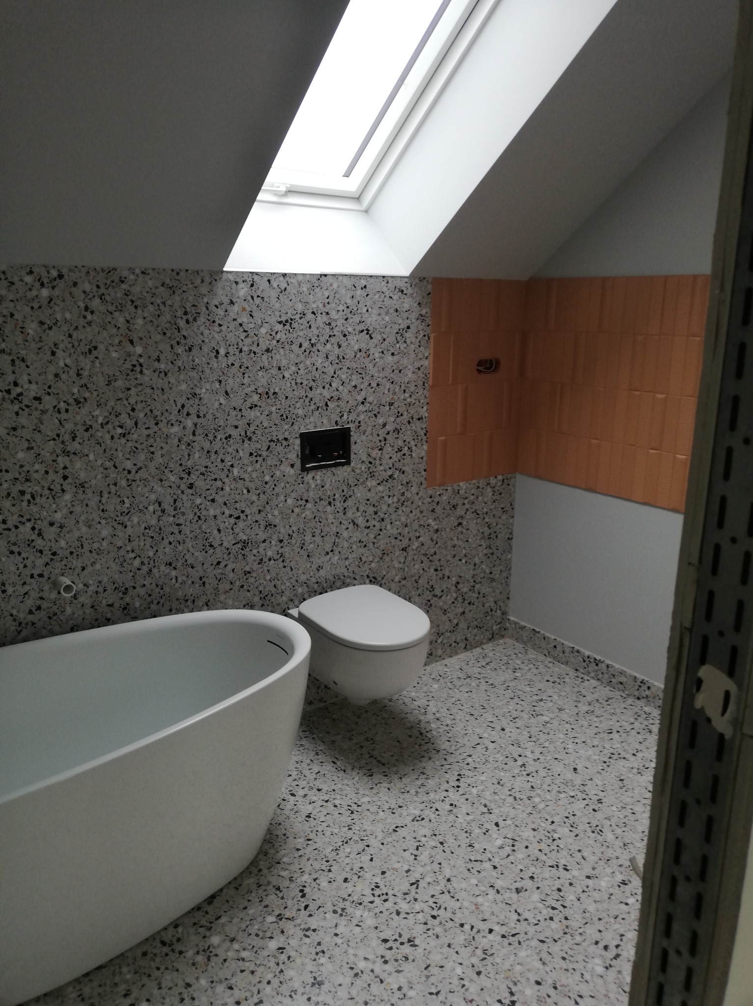 Nowoczesna łazienka na poddaszu z wanną wolnostojącą, toaletą podwieszaną i terazzo na ścianach oraz podłodze. Pomarańczowe płytki na ścianie dodają ciepła.