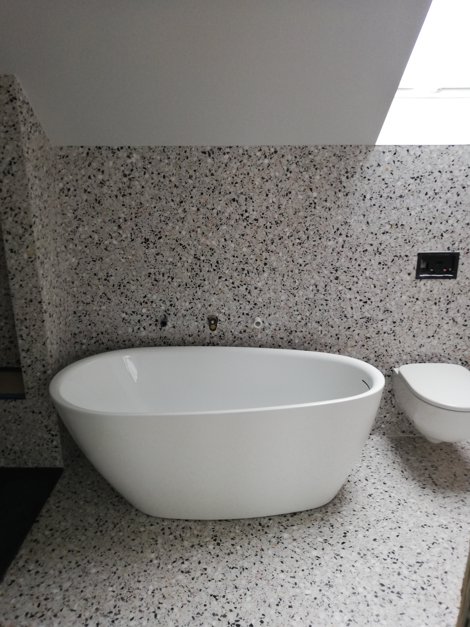 Nowoczesna łazienka z wolnostojącą białą wanną na tle ściany i podłogi wykończonej płytkami lastryko. Minimalistyczny design, górne oświetlenie.