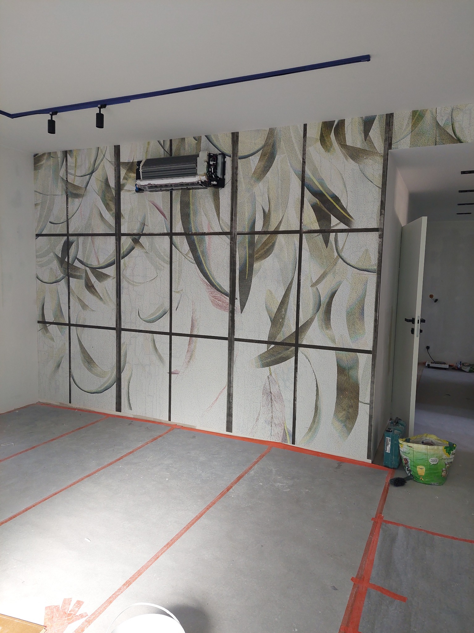 Ściana z dekoracyjną tapetą w motywy roślinne, podzielona ciemnymi listwami. Widoczne oświetlenie sufitowe i fragment klimatyzacji. Podłoga w trakcie prac wykończeniowych.