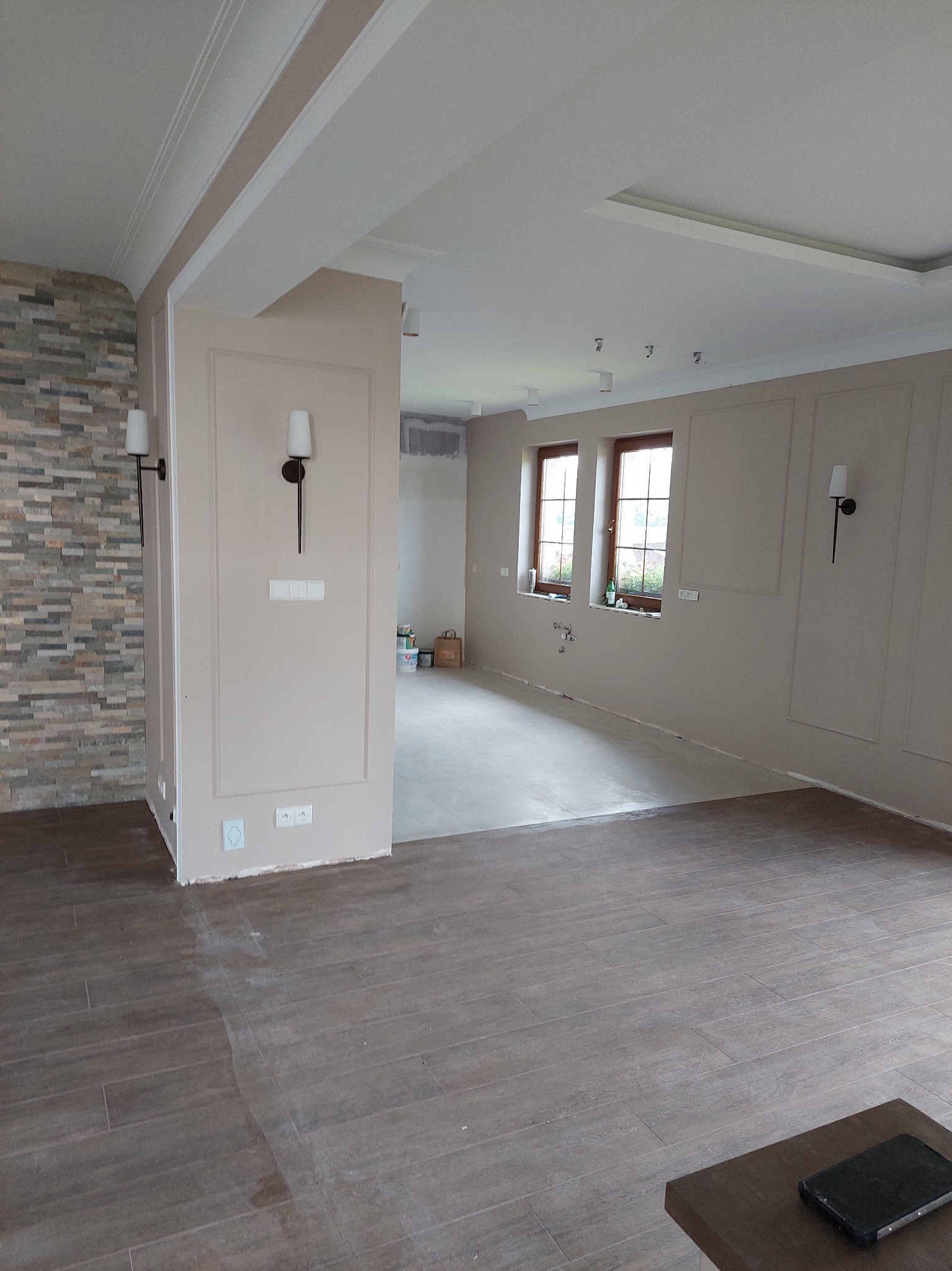 Jasne wnętrze po remoncie: brązowe panele podłogowe, beżowe ściany z dekoracyjnymi listwami, kamienna ściana, nowoczesne lampy ścienne. Widok na dalsze pomieszczenia.