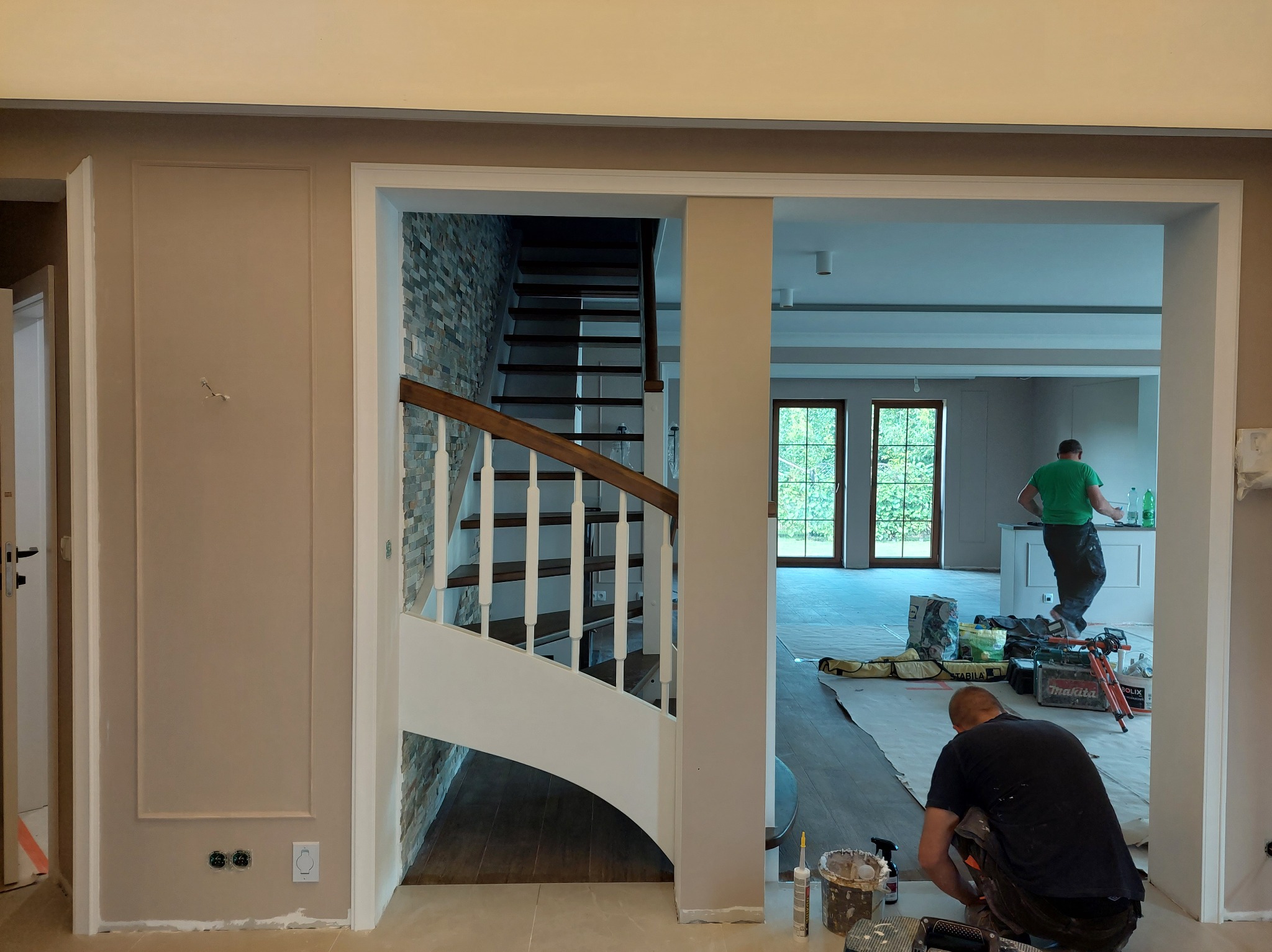 Prace wykończeniowe w domu: widok na schody z kamienną ścianą i otwartą przestrzeń. Dwóch mężczyzn pracuje, widoczne narzędzia i materiały budowlane.