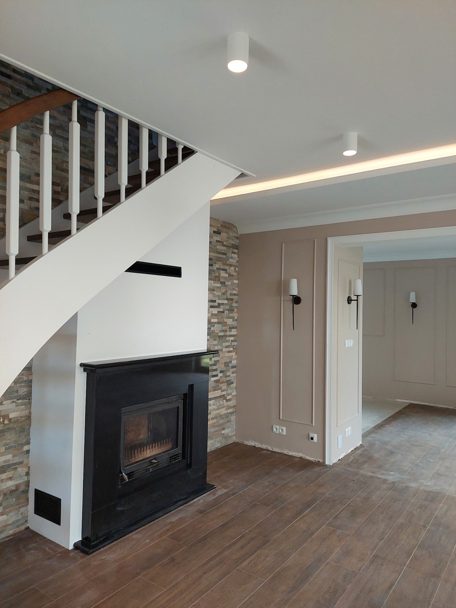 Salon po remoncie: drewnopodobna podłoga, kominek z czarną obudową i kamienną ścianą, białe schody, beżowe ściany z dekoracyjnymi listwami i czarnymi kinkietami.