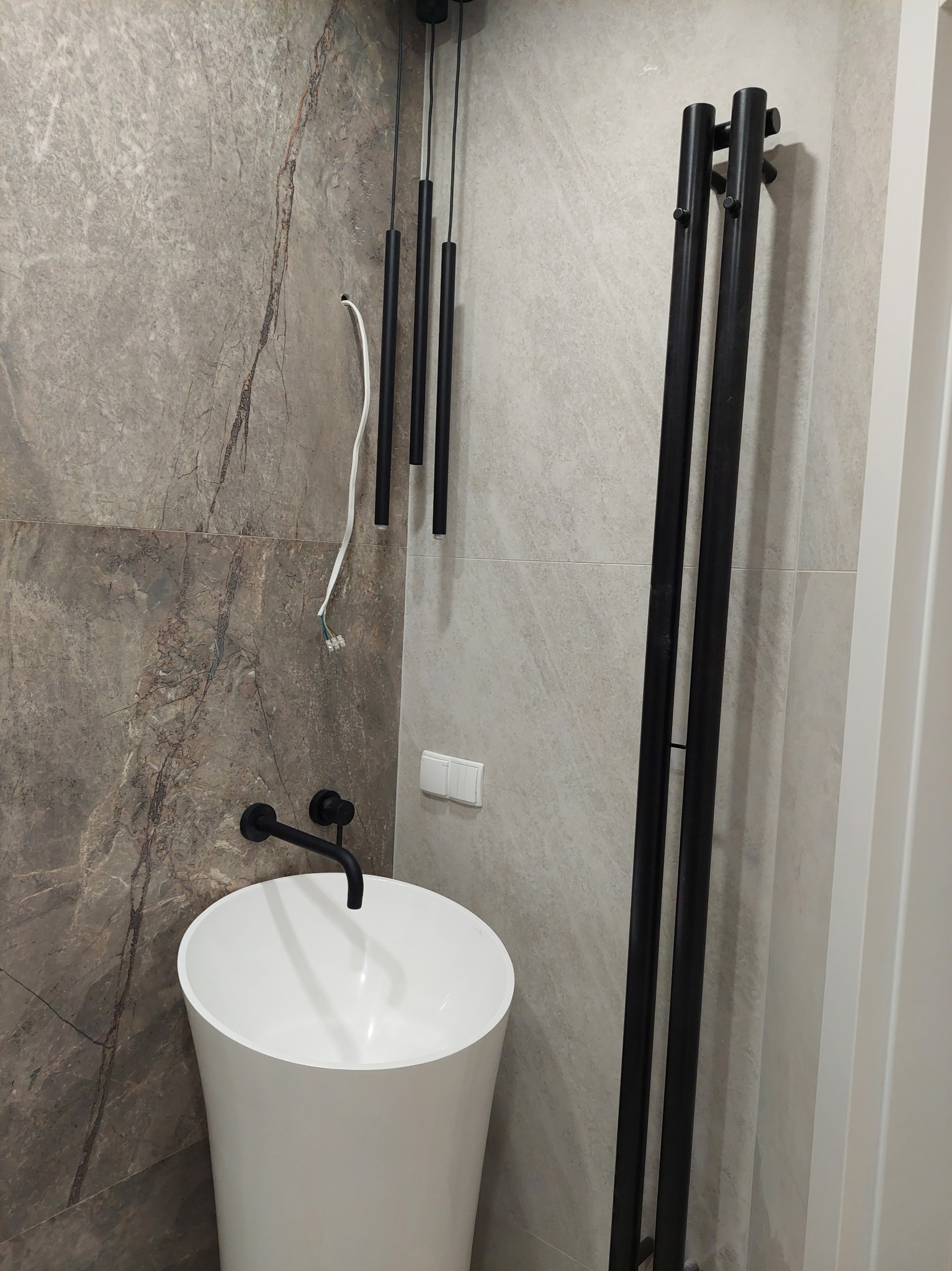 Nowoczesna łazienka w trakcie wykańczania: biała umywalka wolnostojąca, czarny grzejnik drabinkowy i lampy wiszące na tle płytek imitujących marmur.