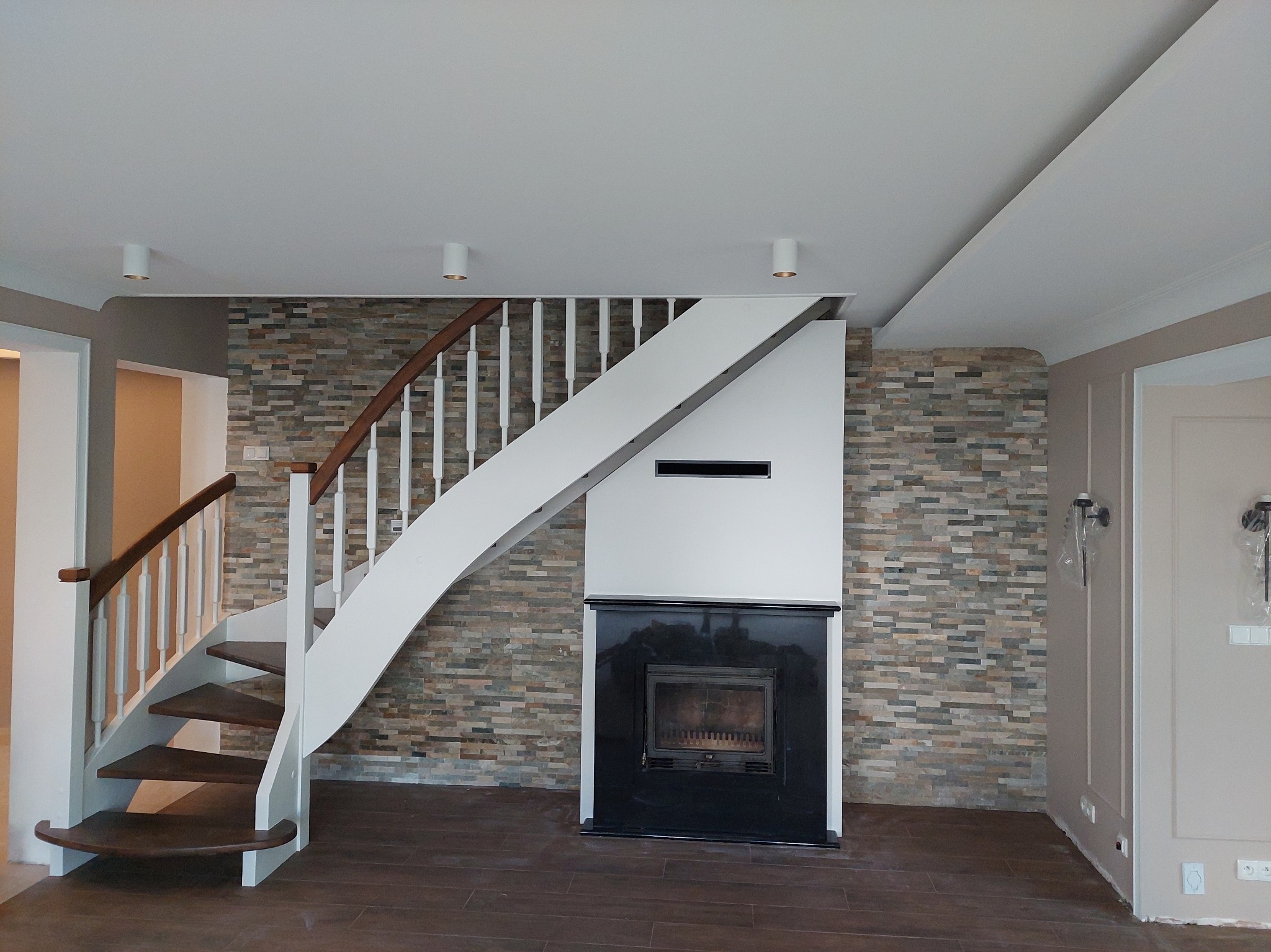 Stylowe wnętrze z drewnianymi schodami o białych balustradach, kamienną ścianą i czarnym kominkiem. Nowoczesne oświetlenie sufitowe. Wykończenie w neutralnych barwach.