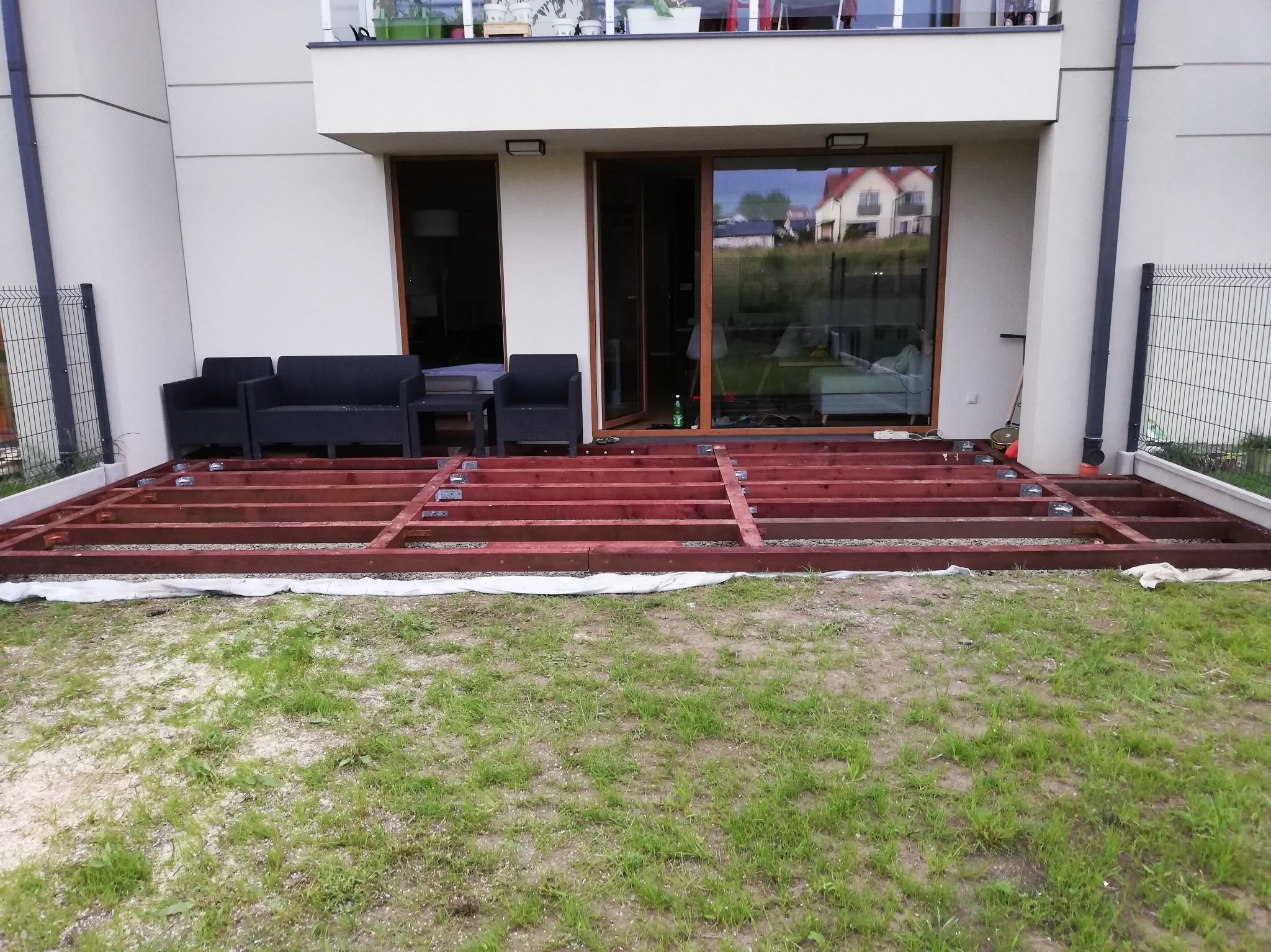 Konstrukcja drewnianego tarasu w trakcie budowy, widok z zewnątrz. Elementy konstrukcyjne zaimpregnowane na czerwono, na trawie. W tle meble ogrodowe i budynek.