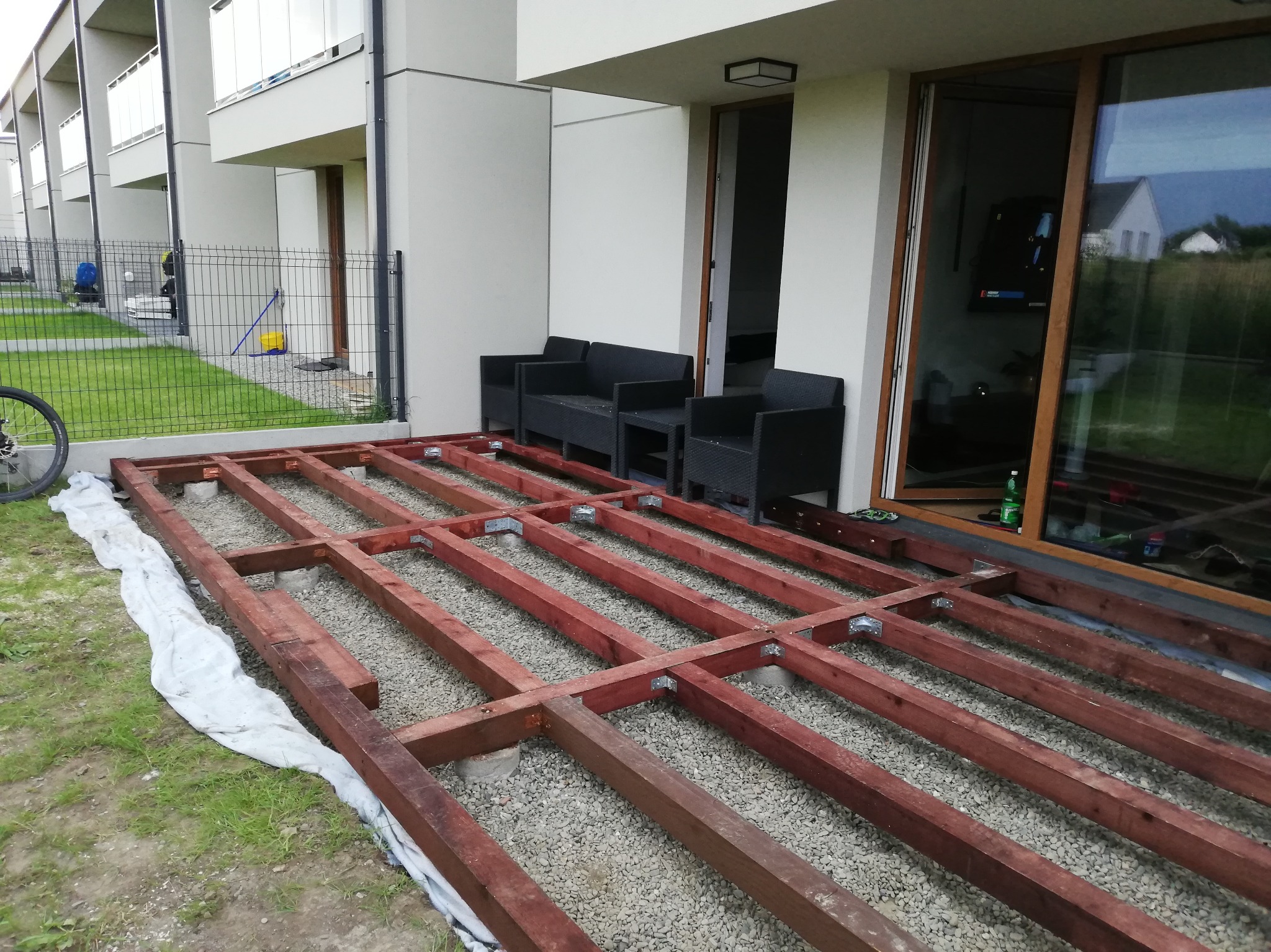 Konstrukcja drewnianego tarasu na żwirowym podłożu, widok z poziomu gruntu. Elementy połączone metalowymi kątownikami, w tle nowoczesny budynek z meblami ogrodowymi.