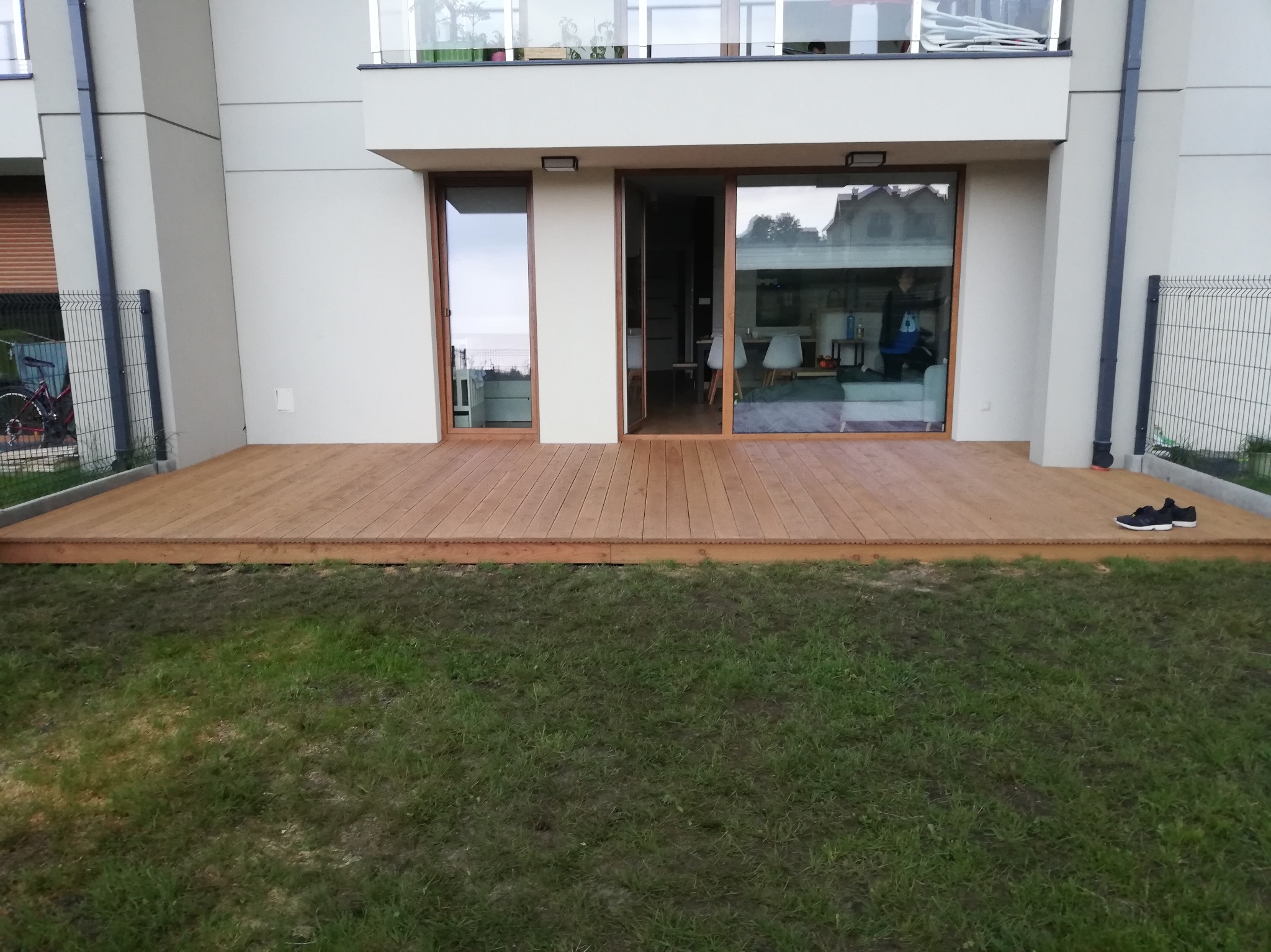 Nowoczesny taras z drewna egzotycznego przy domu z dużymi oknami przesuwnymi. Na trawie leżą buty sportowe. Widok na wnętrze domu.