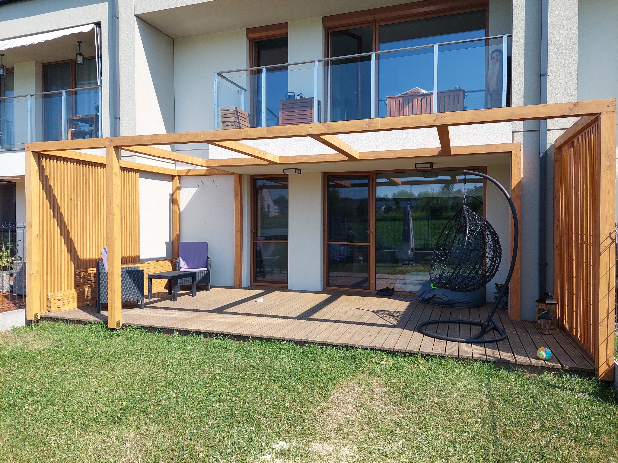 Drewniana pergola z ażurowymi ściankami na tarasie z desek. W tle budynek z balkonami i dużymi oknami. Na tarasie wiszące krzesło i meble ogrodowe.