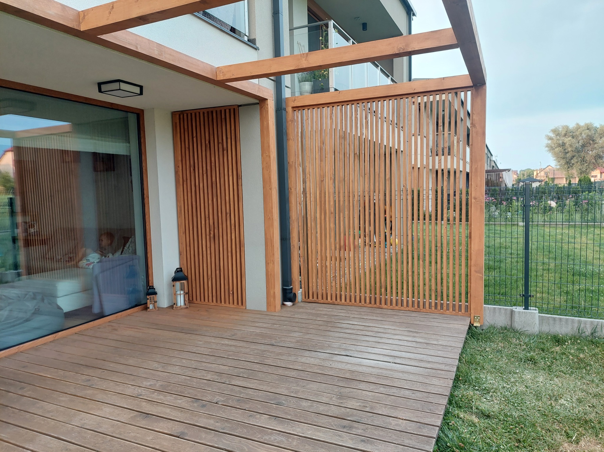 Nowoczesny taras z drewnianą podłogą i ażurową przesłoną z pionowych listew, tworzący strefę relaksu przy domu z dużym oknem.