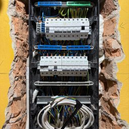 SYLGRE Instalacje Damian Janiszewski - Rozdzielnica elektryczna Schneider w surowej wnęce ściany, widoczne kable, bezpieczniki i listwy. Instalacja w trakcie prac, żółta ściana w tle.