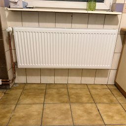 SYLGRE Instalacje Damian Janiszewski - Biały grzejnik panelowy zamontowany na ścianie z beżowych płytek w łazience. Widoczne rury miedziane, termostat i fragment szafki. Ujęcie z perspektywy podłogi.