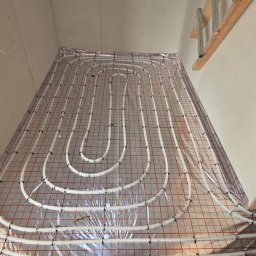 SYLGRE Instalacje Damian Janiszewski - Białe rury ogrzewania podłogowego ułożone na folii z czerwoną siatką, widoczne między drewnianymi elementami konstrukcyjnymi. Część instalacji poza kadrem.