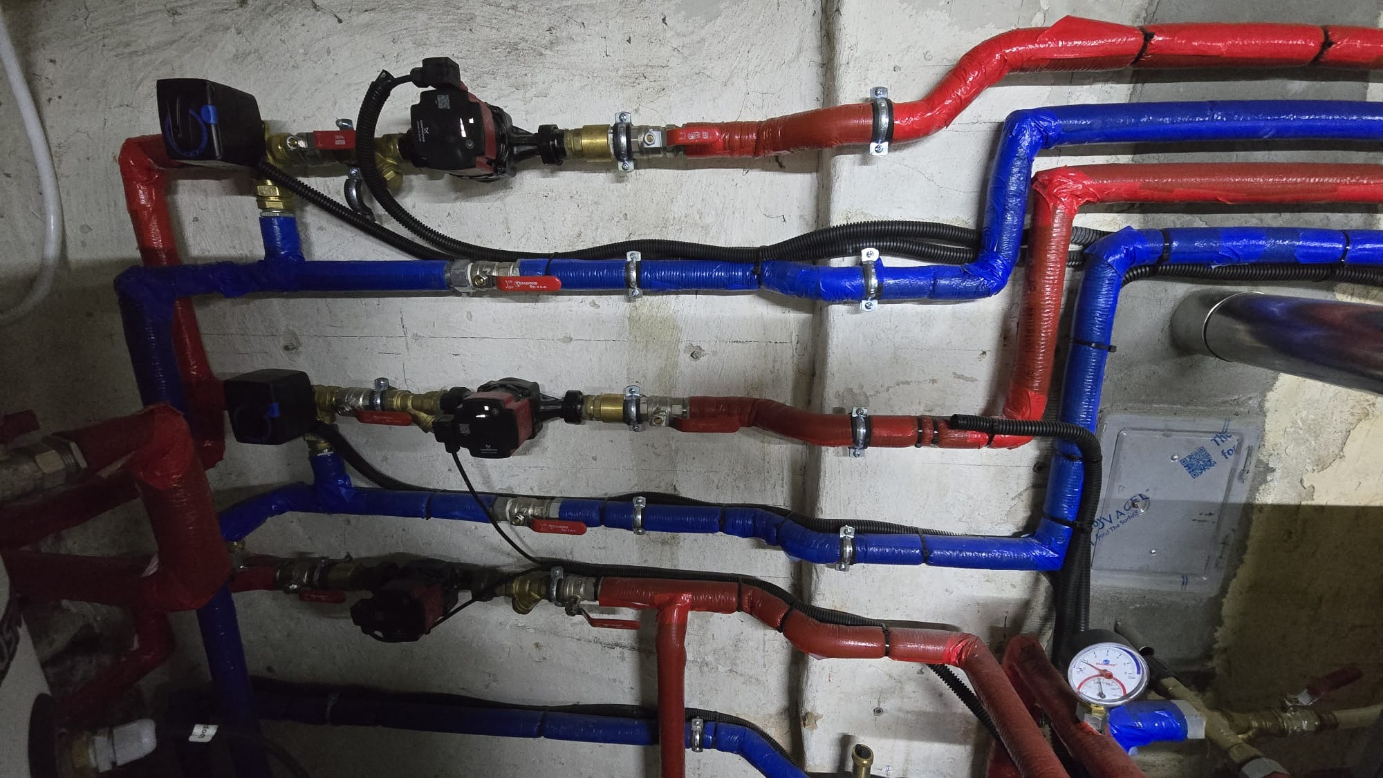 Złożona instalacja hydrauliczna z rurami w izolacji (czerwona i niebieska) oraz pompami obiegowymi, zamontowana na szarej ścianie. Widoczne zawory i manometr.