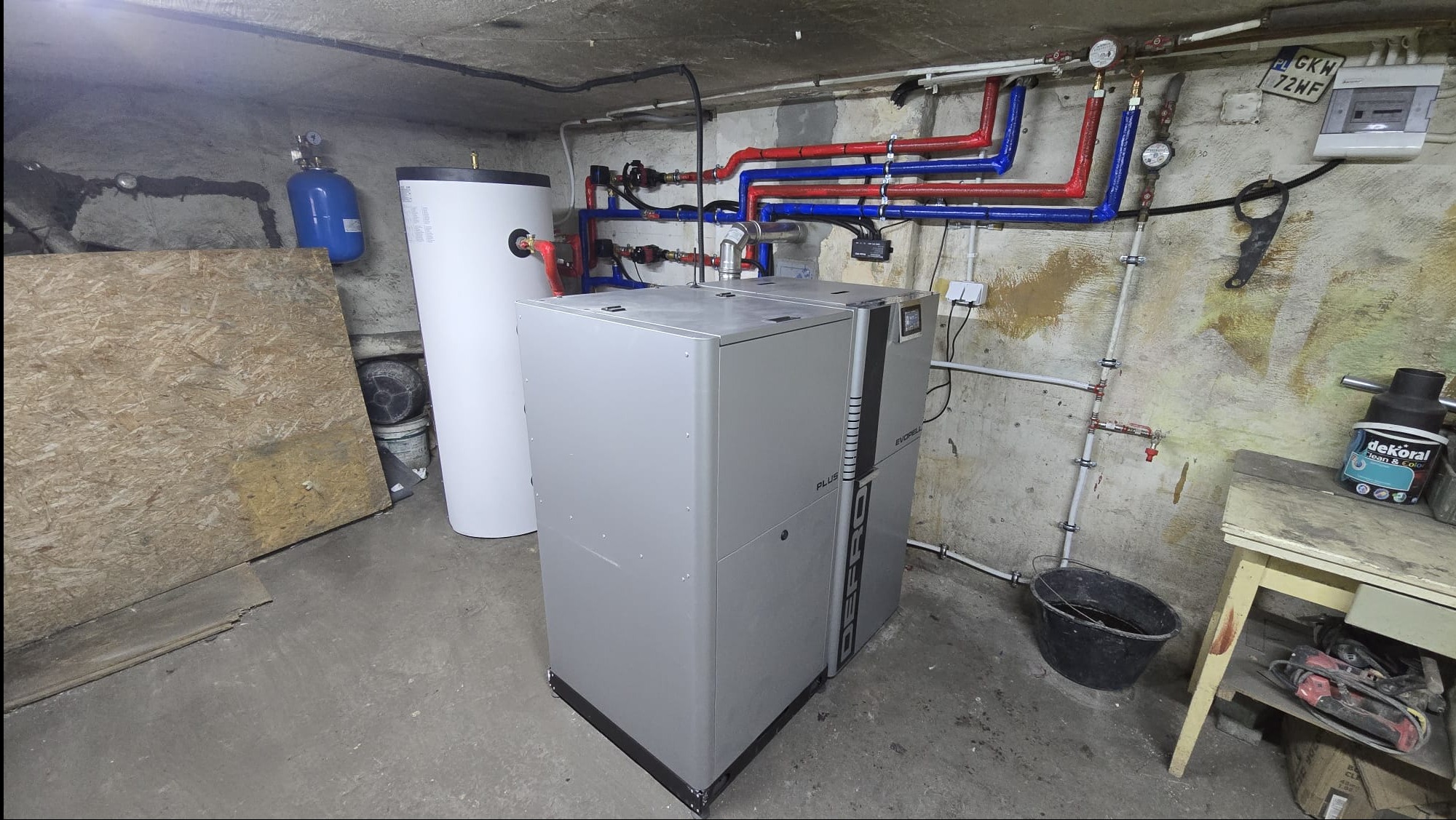 Szary piec Defro Evorell Plus z białym zasobnikiem wody w kotłowni. Widoczne rury CO w kolorach czerwonym i niebieskim oraz osprzęt instalacyjny na ścianie.
