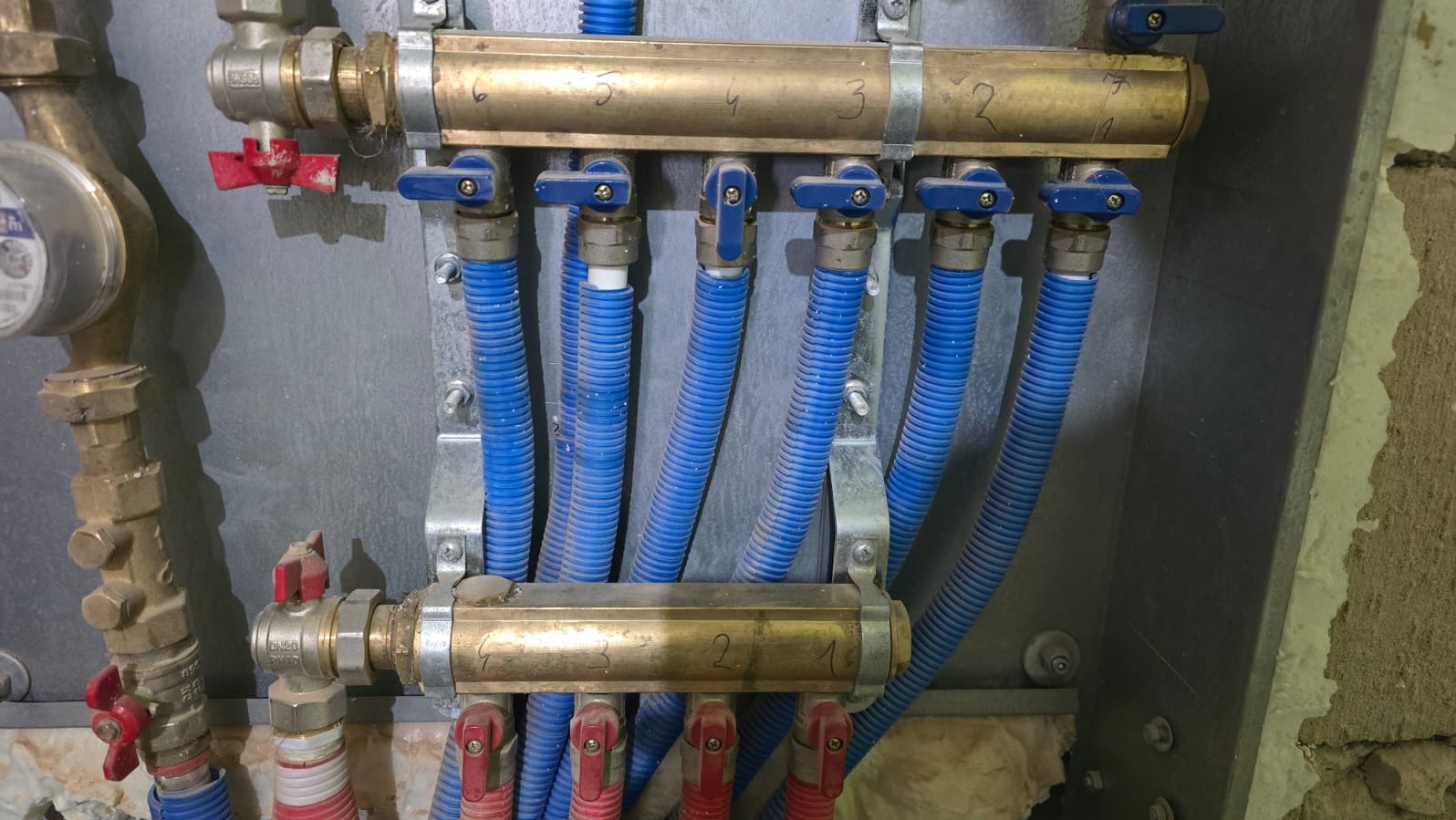 Rozdzielacz instalacji grzewczej z niebieskimi rurami, zamontowany na szarej płycie. Widoczne zawory i numeracja. Częściowo widoczna ściana w tle.