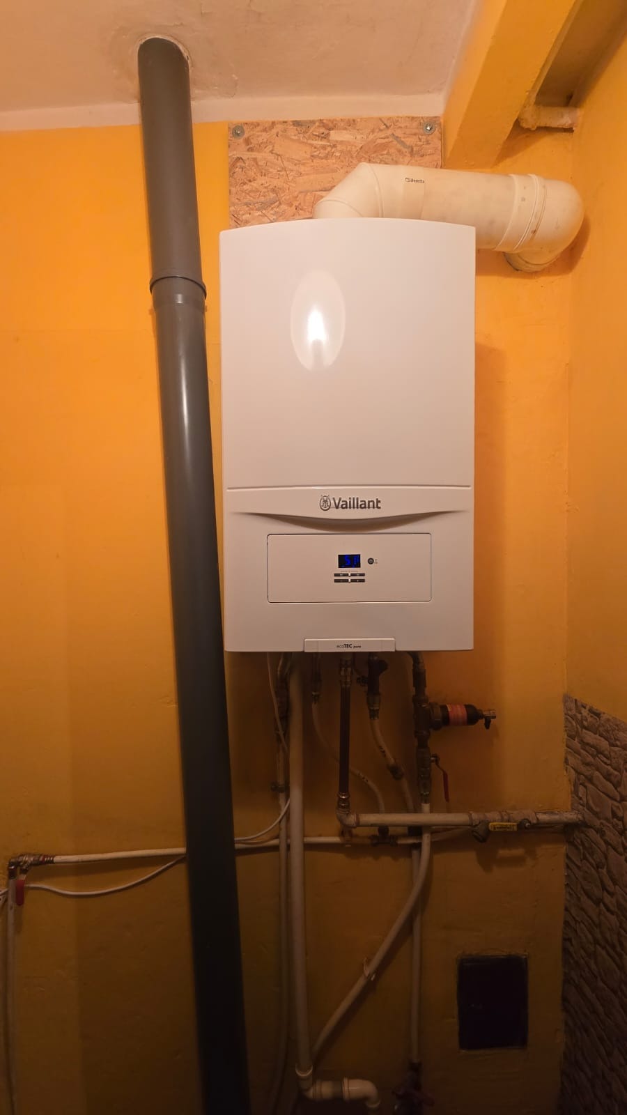 Biały, wiszący piec gazowy Vaillant z podłączonymi rurami i kominem na żółtej ścianie. Widoczne elementy instalacji hydraulicznej i gazowej.