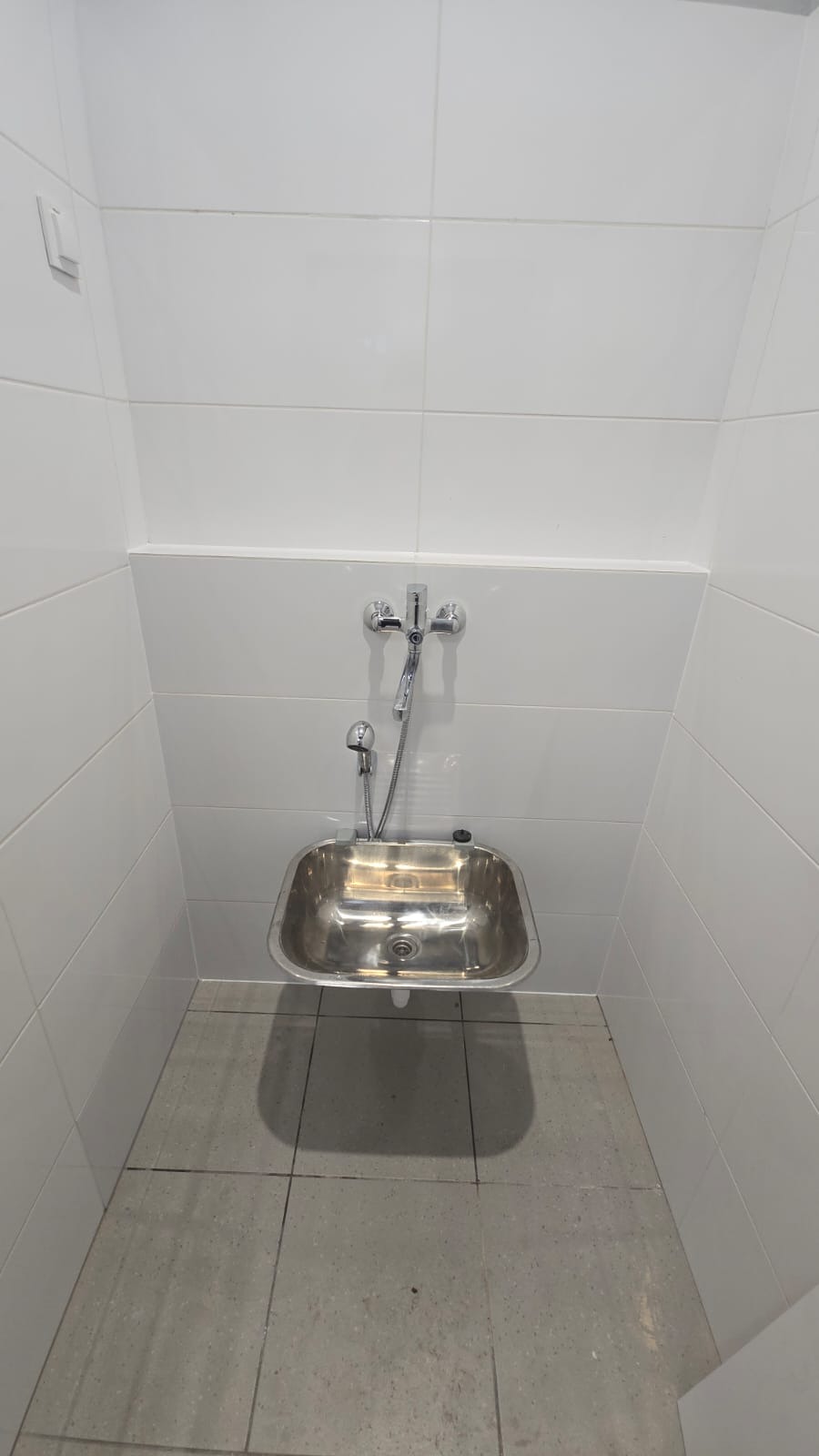 Niewielka, metalowa umywalka ze słuchawką prysznicową zamontowana na białej, kafelkowanej ścianie w małej łazience. Minimalistyczny design, surowe oświetlenie.