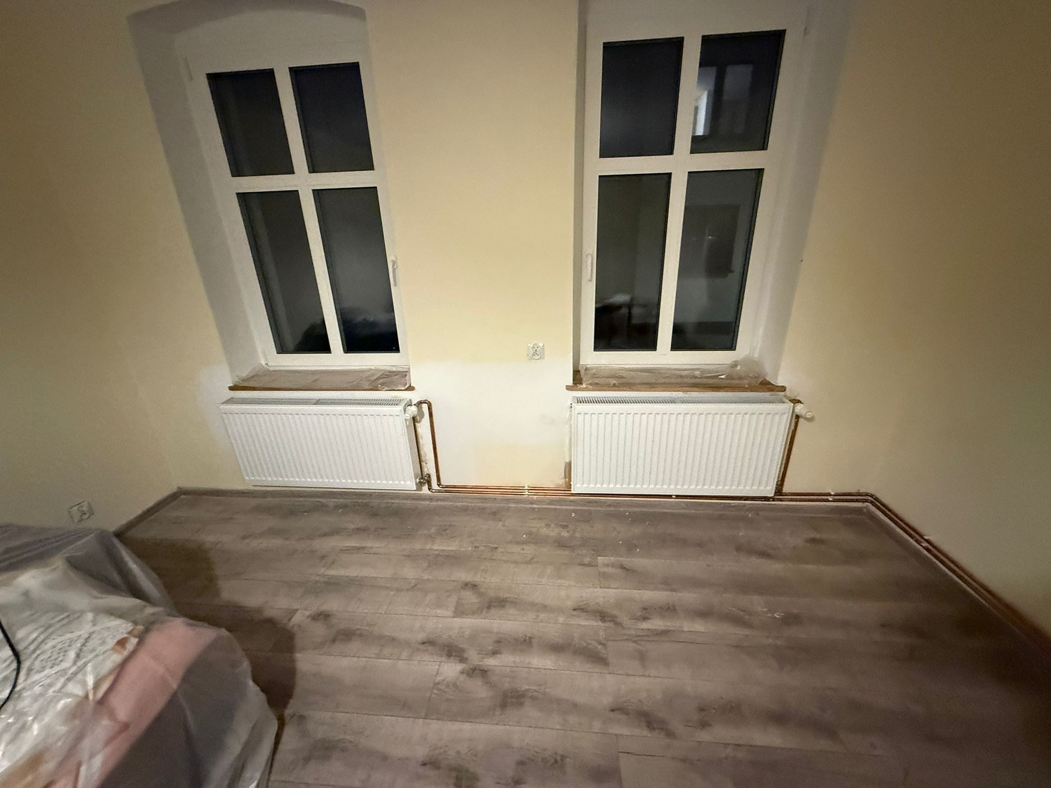 Dwa białe grzejniki pod oknami na tle beżowej ściany i drewnopodobnej podłogi. Widoczne miedziane rury instalacji grzewczej. Część podłogi zakryta folią ochronną.