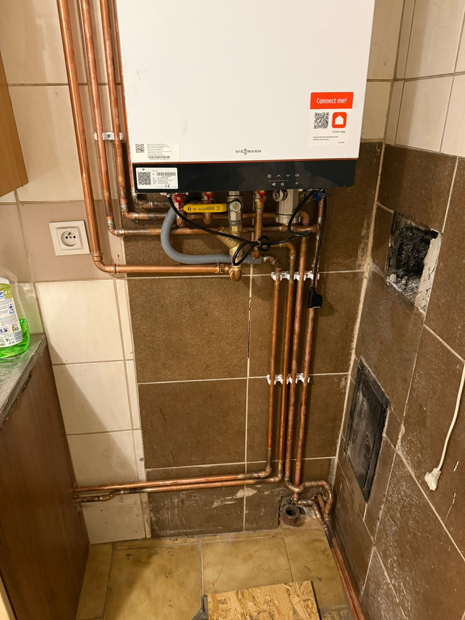 Biały piec gazowy Viessmann z miedzianą instalacją, widoczne rury i zawory. Brązowe płytki na ścianie i podłodze. Gniazdko elektryczne i fragment szafki po lewej.