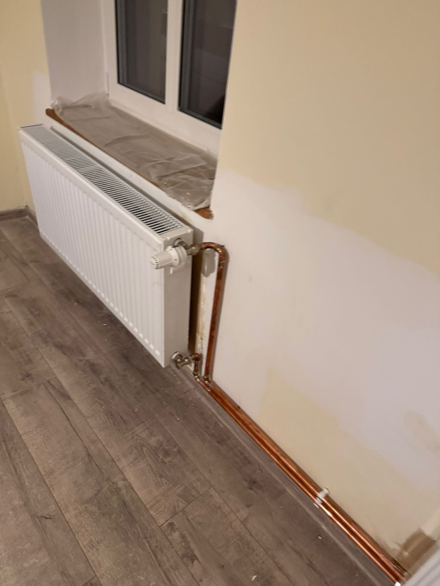 Biały grzejnik żeberkowy pod oknem, z miedzianymi rurami przy ścianie w trakcie montażu. Okno zabezpieczone folią malarską. Podłoga panelowa.