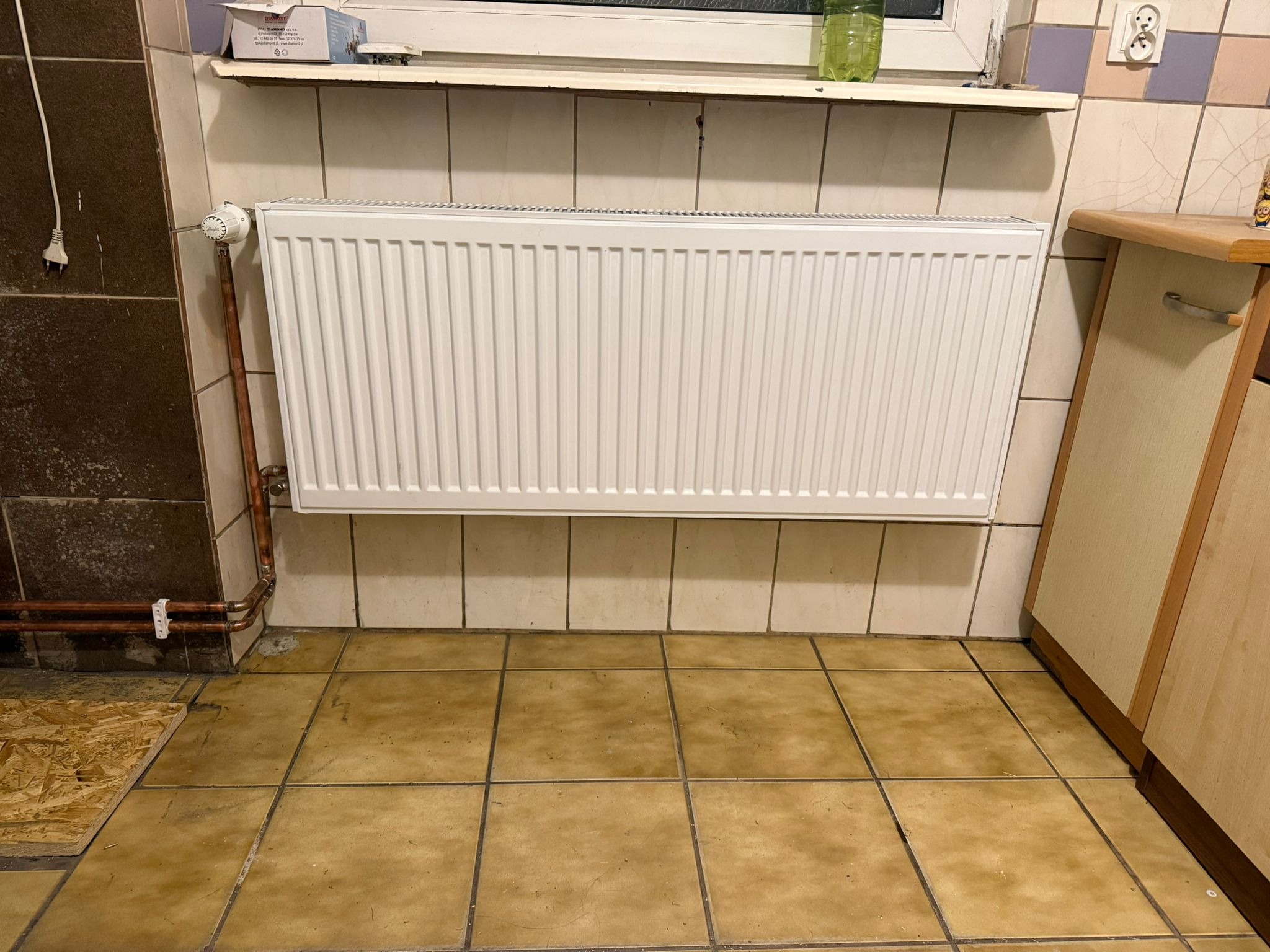 Biały grzejnik panelowy zamontowany na ścianie z beżowych płytek w łazience. Widoczne rury miedziane, termostat i fragment szafki. Ujęcie z perspektywy podłogi.