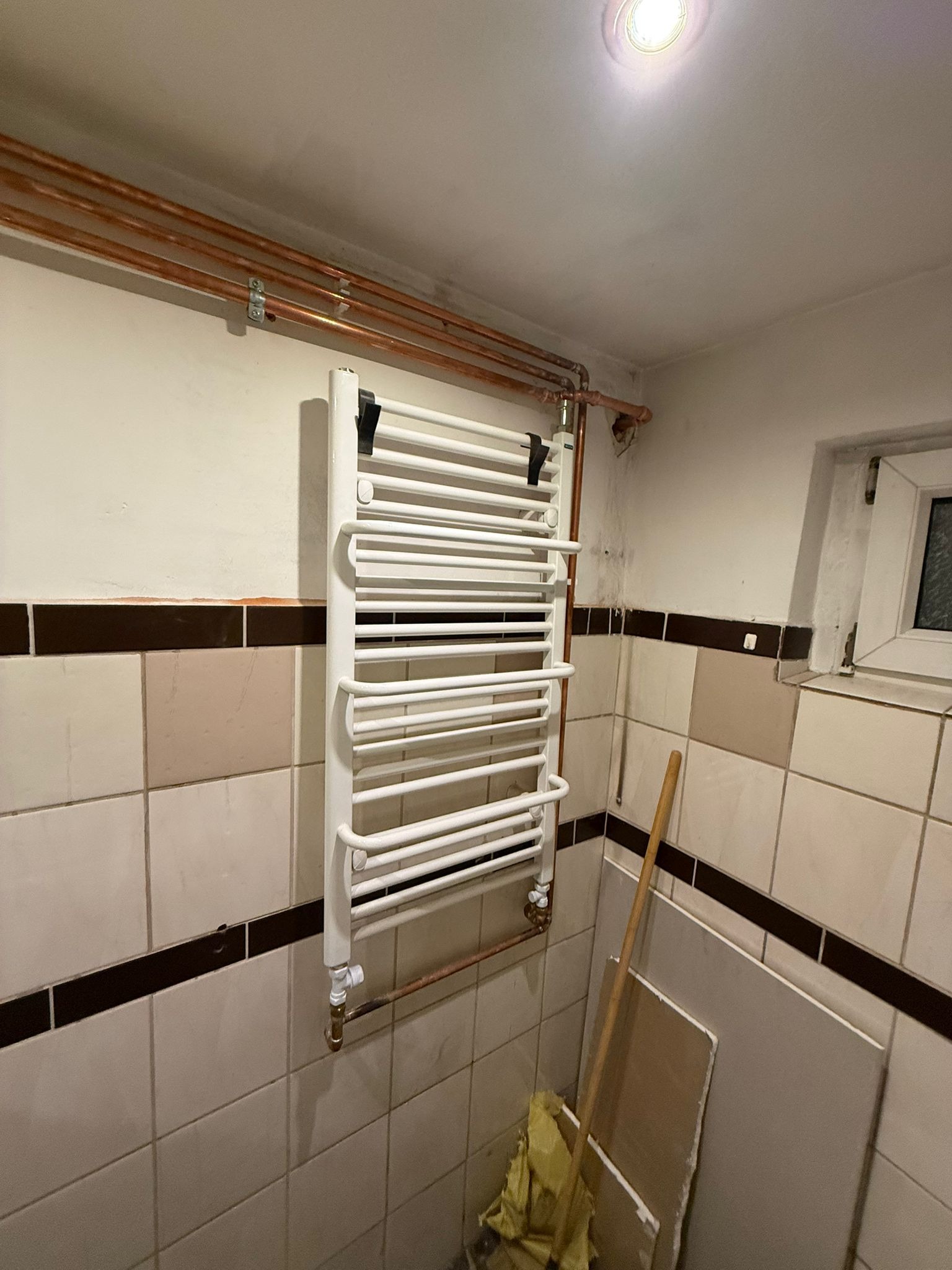 Biały grzejnik łazienkowy drabinkowy zamontowany na ścianie z beżowymi płytkami, widoczne miedziane rury instalacji grzewczej i fragment okna.