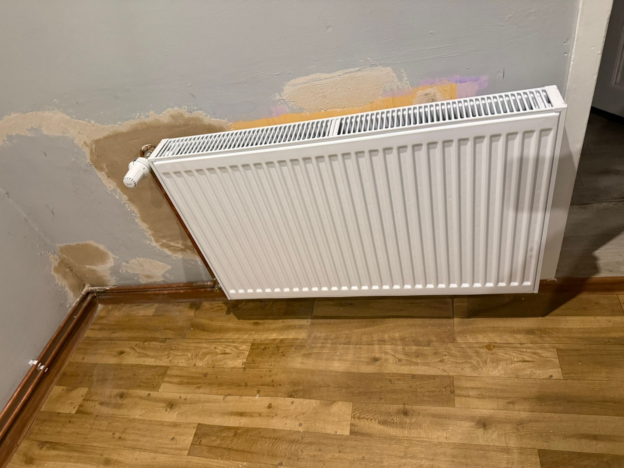 Biały, nowy grzejnik panelowy zamontowany na ścianie z widocznymi śladami po naprawach. Podłoga wyłożona panelami imitującymi drewno. Widoczny zawór termostatyczny.