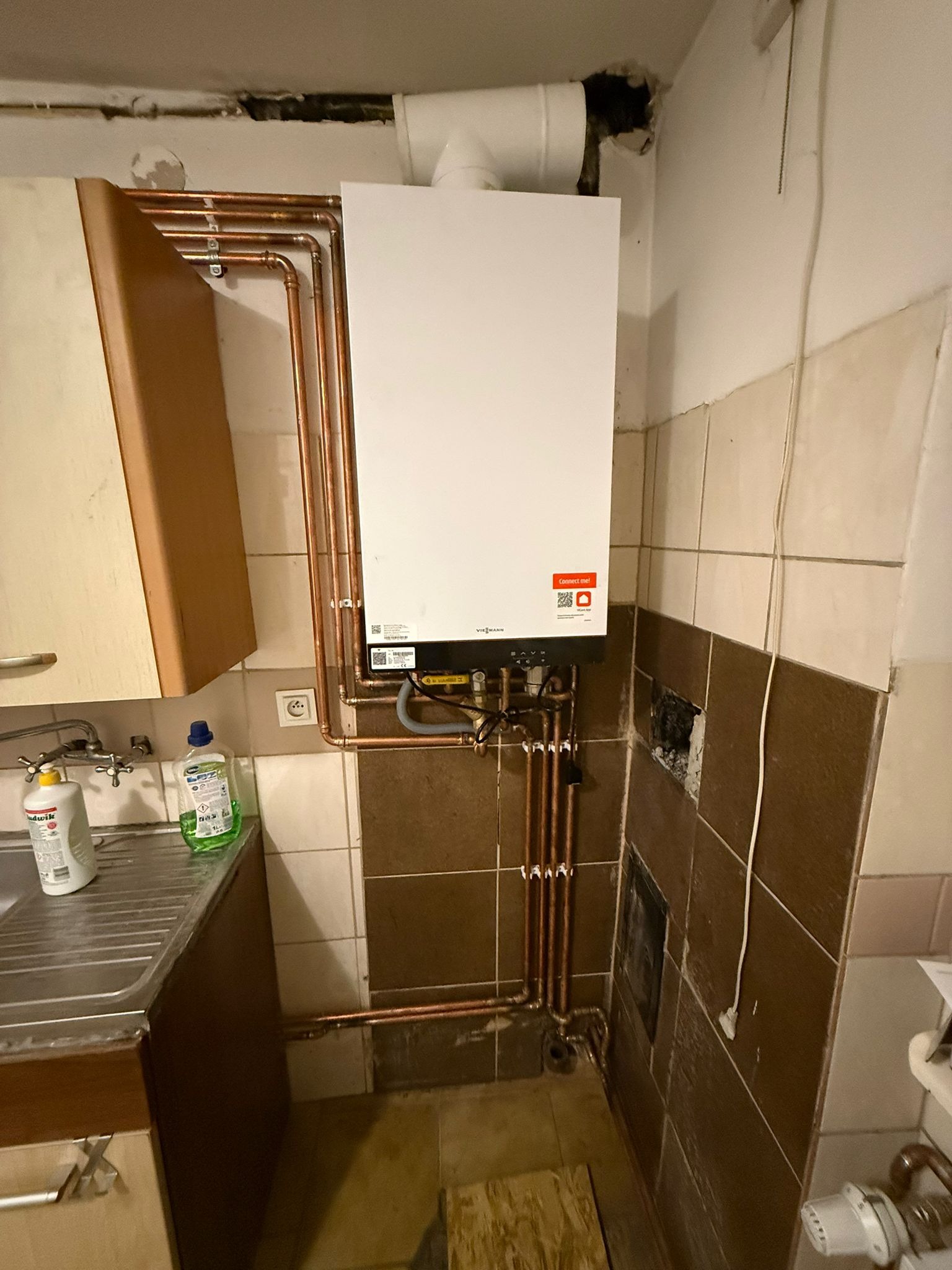 Biały piec gazowy Viessmann zamontowany na brązowo-beżowej ścianie z glazury, z widocznymi miedzianymi rurami instalacji. Obok zlewozmywak z płynami do mycia.