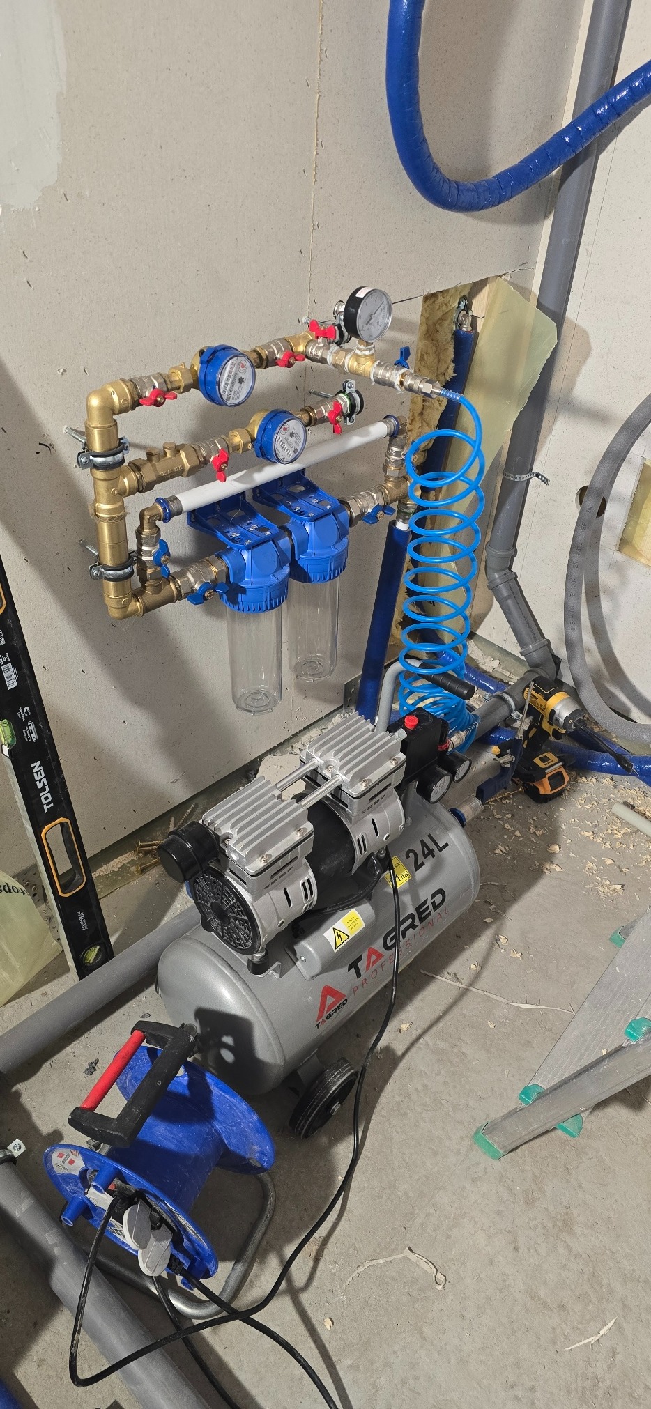 Złożona instalacja wodna z filtrami i manometrami, obok kompresor Tagred 24L, poziomica Tolsen i zwinięty niebieski przewód pneumatyczny. Widoczne rury i narzędzia.