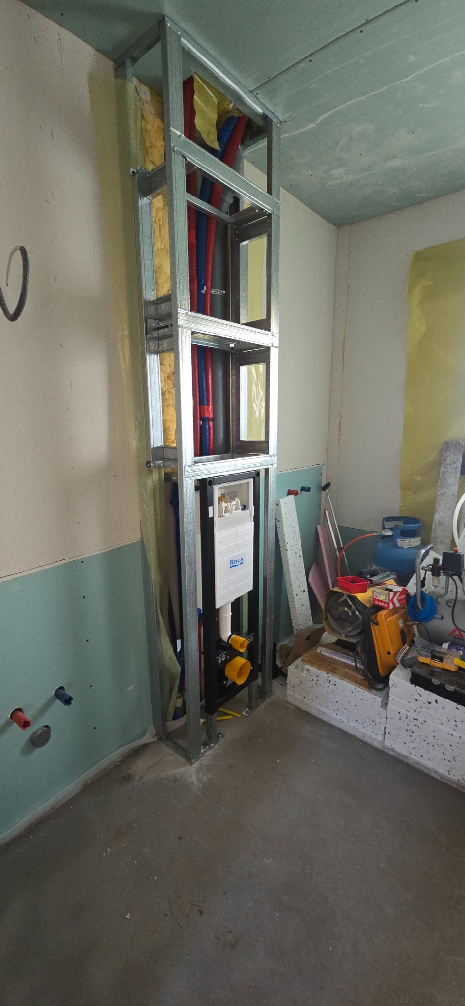 Stelaż podtynkowy WC w trakcie montażu, widoczne rury instalacyjne w kolorach czerwonym i niebieskim, izolacja z wełny mineralnej. W tle narzędzia i materiały budowlane.