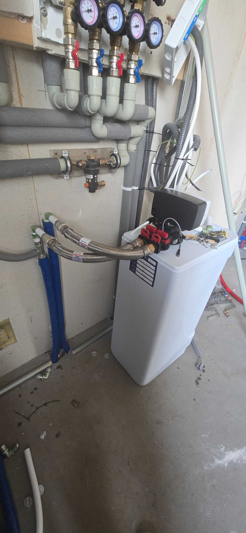 Biały zmiękczacz wody podłączony do instalacji hydraulicznej z rurami w izolacji i manometrami. Widoczne złącza, zawory i przewody. Elementy instalacji elektrycznej.