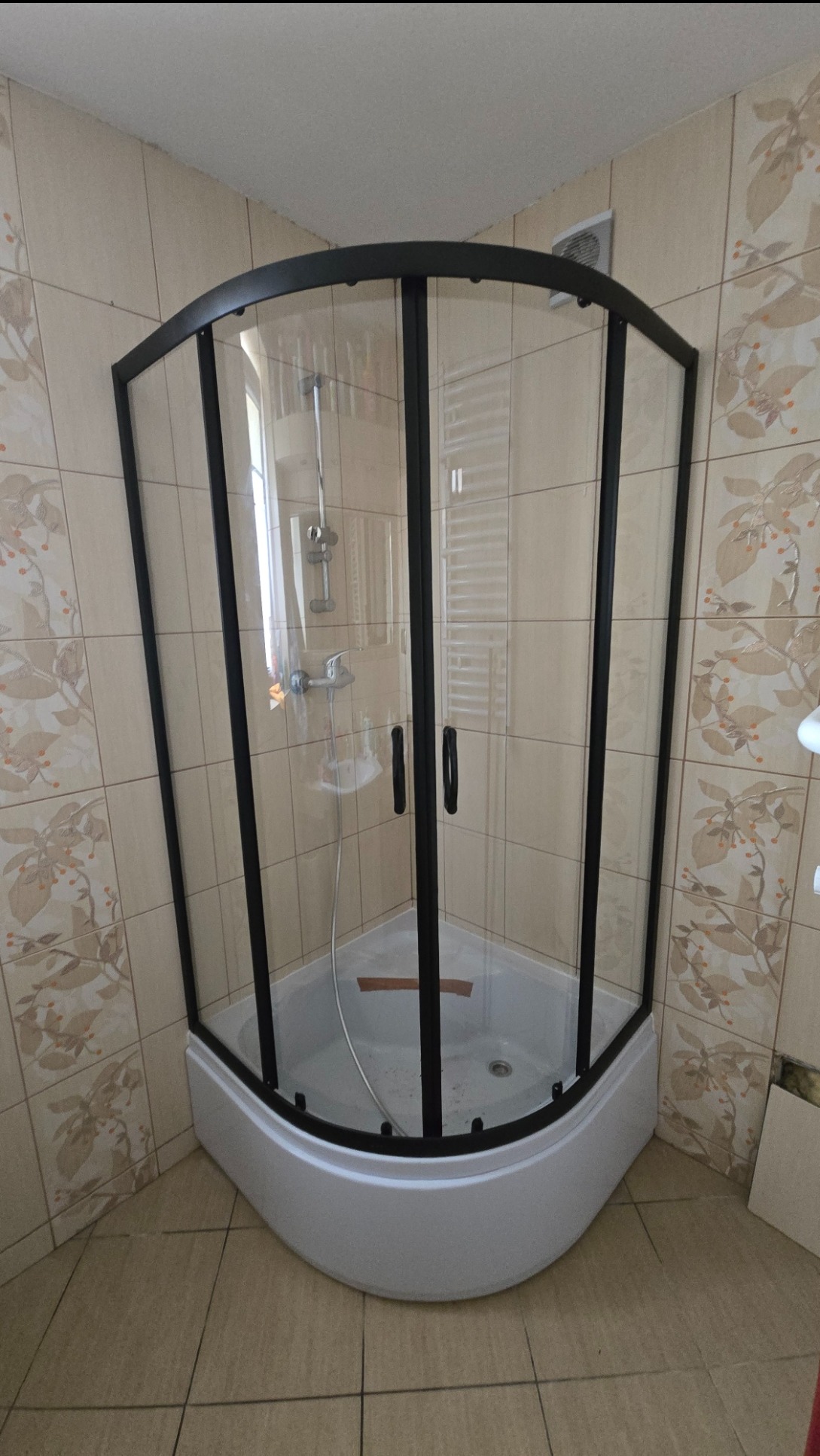 Narożna kabina prysznicowa z czarnymi profilami i brodzikiem, zamontowana w łazience z beżowymi płytkami w kwiatowy wzór. Widoczny odpływ i bateria.