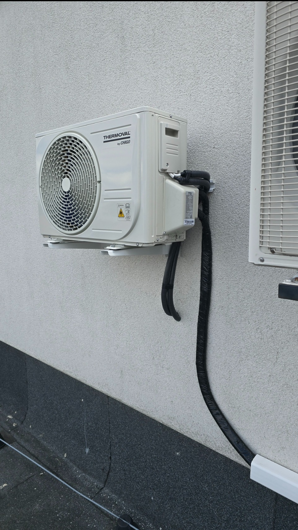 Biały klimatyzator Thermoval by Chigo zamontowany na elewacji budynku, z widocznymi przewodami w czarnej izolacji i białej listwie przypodłogowej.
