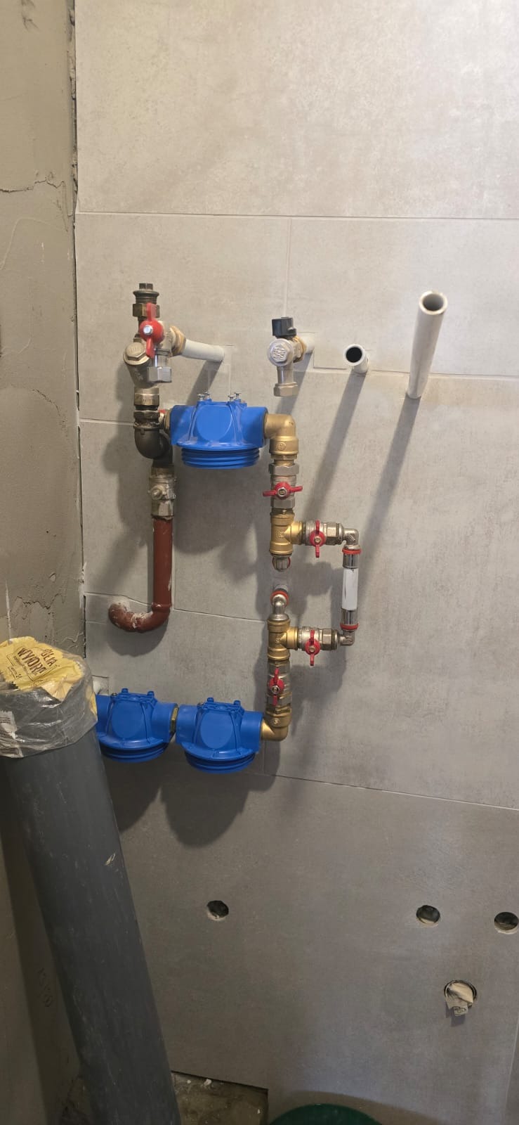 Kompleksowy system filtracji wody z niebieskimi obudowami filtrów, zaworami z czerwonymi uchwytami i rurami miedzianymi na tle szarej ściany z płytkami.
