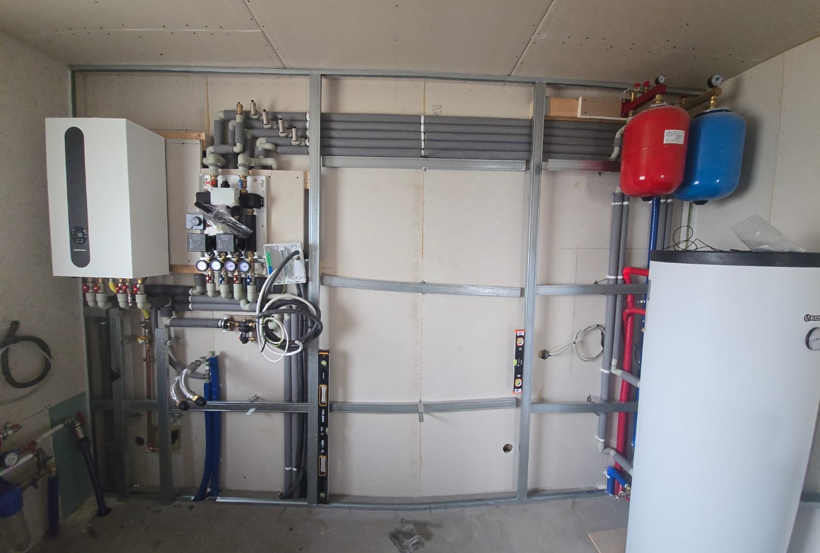 Nowa instalacja grzewcza w trakcie montażu: biały piec, czerwony i niebieski zbiornik, szare rury, metalowa konstrukcja na ścianie z płyt gipsowych. Widoczne poziomice.